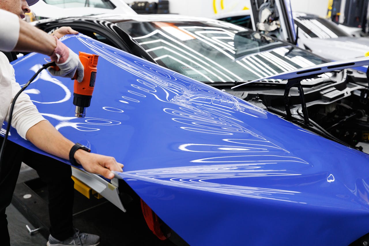 A technician uses a heat gun to carefully apply a blue vinyl wrap to a car in an auto workshop.